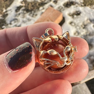 May include: Gold-tone cat charm with a round, orange-brown stone inside. The cat's face is outlined in gold and has a small, gold nose.