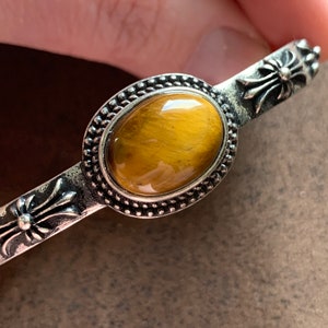 May include: A silver bracelet with a tiger's eye stone set in a silver bezel. The bracelet has a cross design on each end.