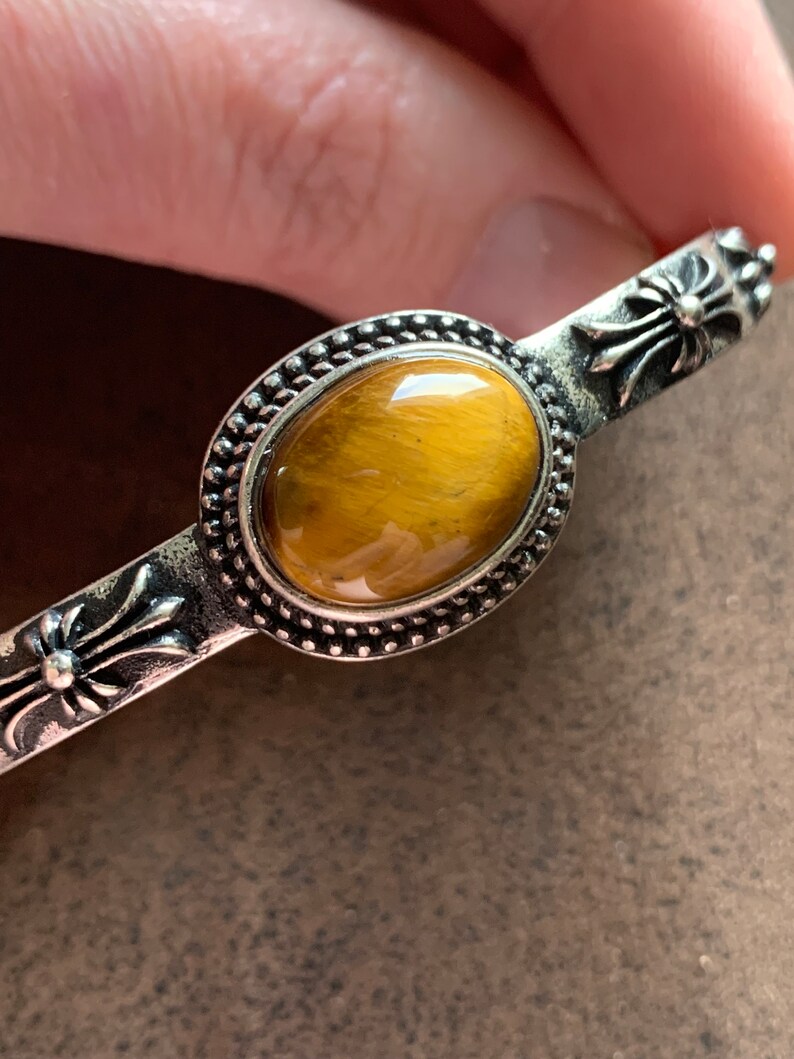 May include: A silver bracelet with a tiger's eye stone set in a silver bezel. The bracelet has a cross design on each end.