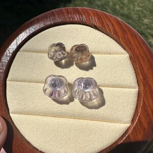 May include: Four clear glass paw print charms, two are a light brown color and two are a light purple color. The charms are displayed on a wooden surface.