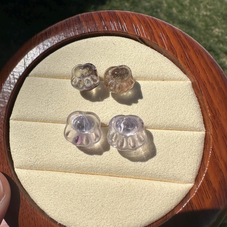 May include: Four clear glass paw print charms, two are a light brown color and two are a light purple color. The charms are displayed on a wooden surface.