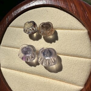 May include: Four glass paw print charms in two different colors. Two are clear with a light purple hue and two are clear with a light brown hue.