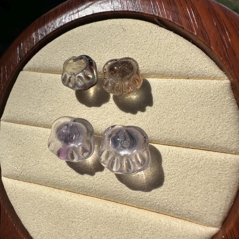 May include: Four glass paw print charms in two different colors. Two are clear with a light purple hue and two are clear with a light brown hue.