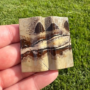 May include: A rectangular piece of polished landscape jasper featuring a distinct V-shaped groove down the center. The stone displays a cream-colored base with horizontal bands of dark brown and black mineral inclusions and dendritic patterns.