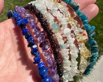 May include: A set of seven gemstone bracelets, each featuring a different color and type of stone. The bracelets are made of small, irregular-shaped stones and are arranged in a rainbow order, starting with blue lapis lazuli, then amethyst, garnet, aquamarine, white quartz, citrine, and teal apatite.