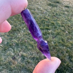May include: A polished, elongated amethyst crystal pendant with a silver-colored loop at the top. The purple crystal is held up against a blurred green background. The pendant is approximately 3 inches long.