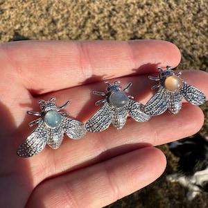 May include: Three silver bee brooches with different colored gemstone accents. The bees have detailed wings and bodies. The gemstones are a light blue, a gray, and a light brown.