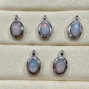 May include: Five oval-shaped pendants featuring iridescent white opals set in polished silver-toned metal frames. Each pendant has a small integrated bail at the top for attachment, showcasing a smooth, rounded cabochon stone with subtle flashes of color.