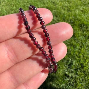 May include: Two strands of a beaded bracelet featuring small, round, deep red garnet stones. The bracelet is held in a hand against a backdrop of green grass, showcasing the jewelry's color and texture.