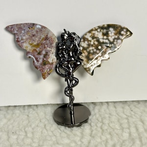 May include: A decorative sculpture featuring a black metal silhouette of two figures embracing, with gemstone wings. The wings are made of polished stone in shades of brown, white, and yellow. The sculpture is mounted on a round, black base.