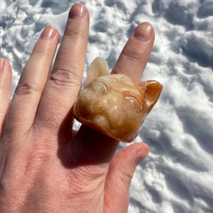 May include: A carved cat head ring made of orange and white agate. The ring is worn on a person's finger.
