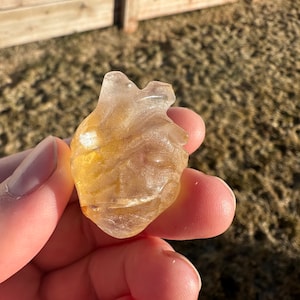 May include: A heart-shaped clear quartz crystal with a golden hue. The crystal is smooth and polished, with a slight sparkle.