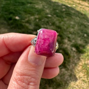 May include: A silver ring with a large, rectangular, pink gemstone. The gemstone has a swirling pattern and is set in a silver band with decorative details.