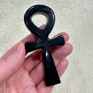 May include: A polished black ankh pendant. The ankh, a cross with a loop at the top, is held in a hand. The pendant has a smooth, glossy finish and is a symbol of life and immortality.