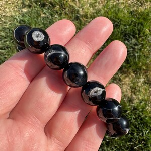 May include: A black beaded bracelet with white and gray inclusions. The bracelet is made of polished, round beads. The beads have a glossy finish and are strung together. The bracelet is held in a hand.
