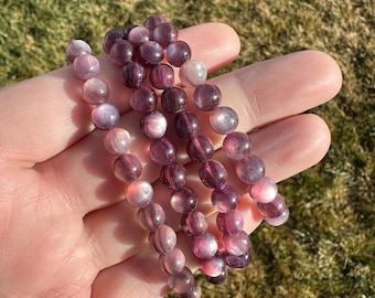 May include: Several strands of purple and pink gemstone bead bracelets. The beads are round and have a polished, translucent appearance. The bracelets are held in a hand against a background of green grass.