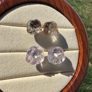 May include: Four clear glass paw print charms, two are a light purple color and two are a smoky brown color. The charms are displayed on a white velvet background.