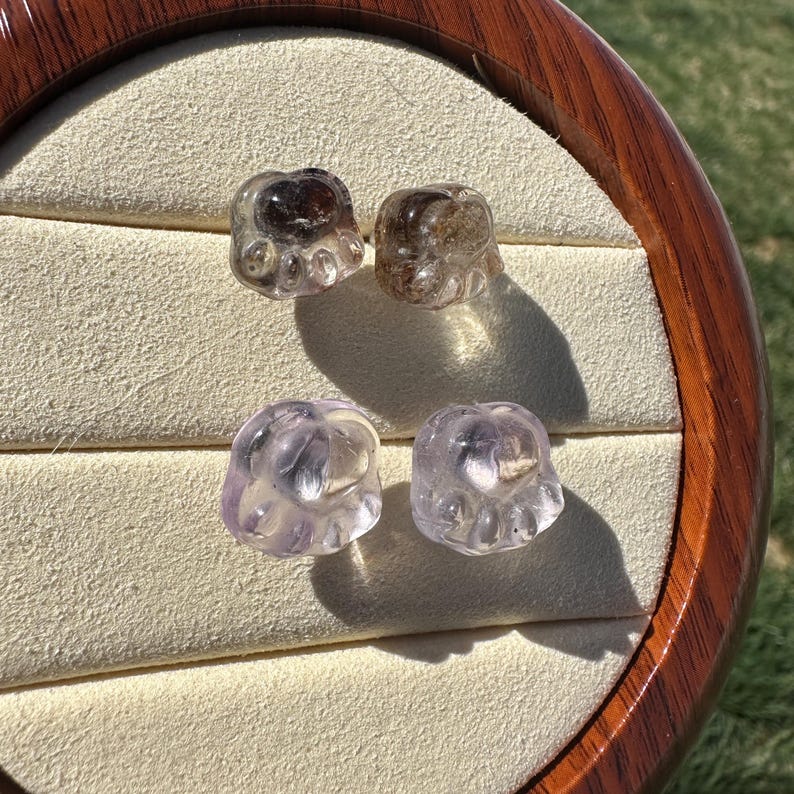 May include: Four clear glass paw print charms, two are a light purple color and two are a smoky brown color. The charms are displayed on a white velvet background.