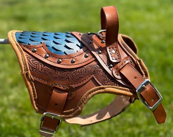 May include: A brown leather harness featuring a blue scale-patterned panel, intricate embossed knotwork detailing, silver-toned metal buckles, and rivets. The harness includes adjustable straps and a top handle, constructed with thick leather edges.