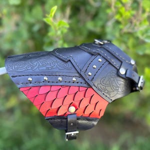 May include: A black leather bracer with intricate embossed designs and silver rivets. The lower portion features a red, scale-like pattern. Adjustable straps and buckles secure the bracer, suggesting a medieval or fantasy theme.