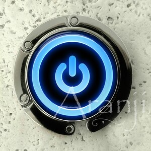 May include: A silver-colored purse hook with a blue illuminated power button design. The hook is circular with a black center and a bright blue power symbol. The background is a textured, light gray surface.
