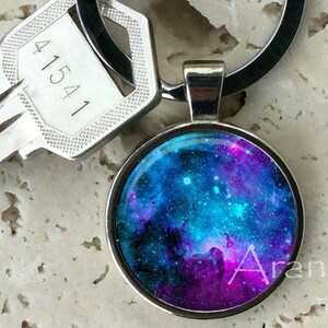 May include: A silver key chain with a round, glass charm featuring a vibrant blue and purple galaxy print. The charm has a silver rim and is attached to a silver ring.