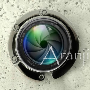 May include: A silver-colored metal keychain shaped like a camera lens. The lens has a black and silver design with the text "ZOOM LENS 70-300 mm 1:2.8 IS USM  ø 77 mm MADE IN JAPAN" printed around the edge.