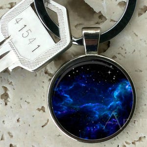 May include: A silver-toned keychain with a round pendant featuring a vibrant blue and purple nebula design with white stars. A silver key with the number "41541" is also attached to the keychain.