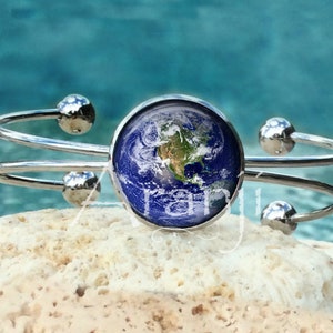 May include: A silver bangle bracelet with a round, blue and green Earth globe charm. The bracelet is made of two bands of metal that wrap around the wrist.