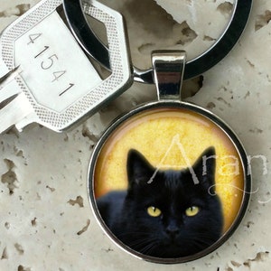 May include: A black cat with yellow eyes is featured on a round, silver keychain. The cat is looking directly at the camera. The keychain is attached to a silver key ring.