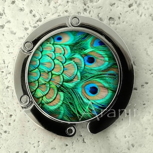 May include: A silver-toned purse hook with a round, colorful design featuring a peacock's tail feathers. The feathers are green, blue, and brown with a pattern of eyes.
