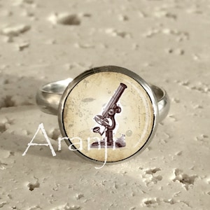 May include: A silver ring with a round, cream-colored cameo featuring a vintage illustration of a microscope.