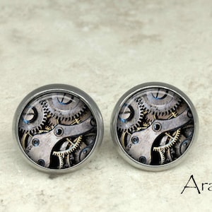 May include: A pair of silver stud earrings with a round, clear glass cabochon featuring a detailed image of a clockwork mechanism with gears and cogs.