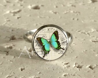May include: A silver ring with a round, white cameo featuring a blue and green butterfly on a vintage postcard. The postcard has the text "Post Card" and "Correspondence" on it.