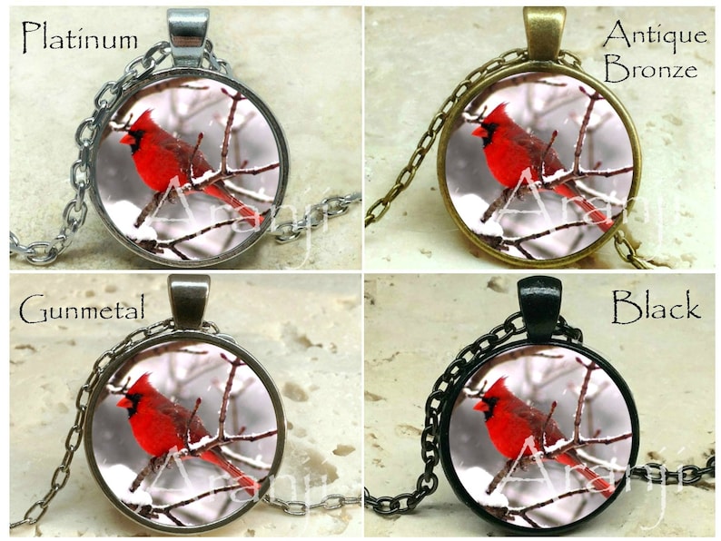 May include: A necklace with a round pendant featuring a red cardinal perched on a snowy branch. The pendant is available in platinum, antique bronze, gunmetal, and black.