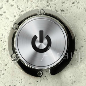 May include: A silver-colored metal bag hook with a circular design featuring a brushed metal finish and a black power button symbol.
