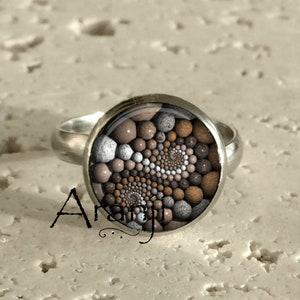 May include: A silver ring with a round, brown and white patterned glass stone. The stone features a spiral design of small, round shapes.