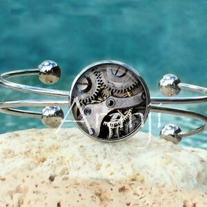 May include: A silver bangle bracelet with a round, clear glass cabochon featuring a vintage clockwork design. The cabochon is set in a silver bezel.