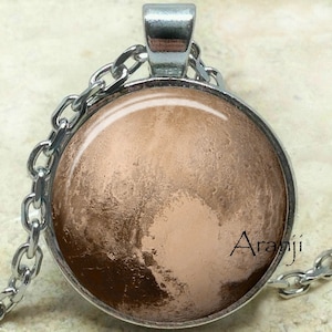 May include: A silver chain necklace with a round glass pendant featuring a brown and beige image of the planet Pluto. The pendant has a silver rim and the text "Aranji" is visible in the lower right corner of the image.