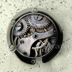 May include: A silver-toned metal keychain with a round, clear plastic insert featuring a detailed image of a clockwork mechanism with gears and cogs.