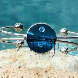 May include: A silver bangle bracelet with a round glass charm featuring a blue and white image of a full moon over a lake and mountains.