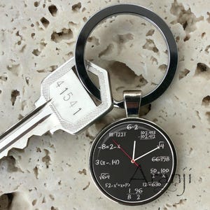 May include: A silver key chain with a black round charm featuring mathematical equations and symbols. The charm has a silver ring attached to it. The key chain is attached to a silver key with the number 41541 engraved on it.