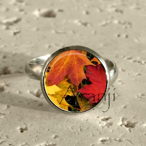 May include: A silver ring with a round, domed centerpiece featuring a vibrant autumn leaf design. The leaves display shades of red, orange, and yellow, set against a dark background. The ring is set on a textured, off-white surface.
