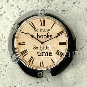 May include: A silver-toned, round, clock-shaped bag charm with a cream-colored face. The clock face has Roman numerals and black hands. The text on the clock face reads "So many books, So little time".