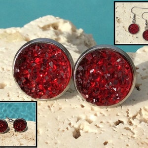 May include: A pair of red glitter stud earrings with silver settings. The earrings are shown on a white textured surface.