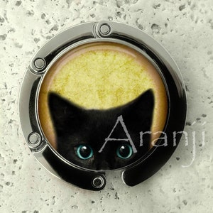 May include: A silver bag clip with a round, clear plastic insert featuring a black cat with bright blue eyes. The cat is looking directly at the viewer. The background is a yellow color.