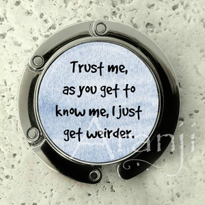 May include: A silver-toned metal bag hook with a blue background and the text "Trust me, as you get to know me, I just get weirder."