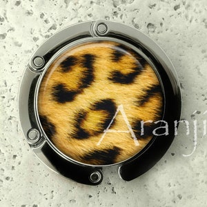 May include: A silver-toned purse hook with a black accent. The center features a round design with a leopard print pattern in shades of brown and black. The word "Aranji" is printed on the design.