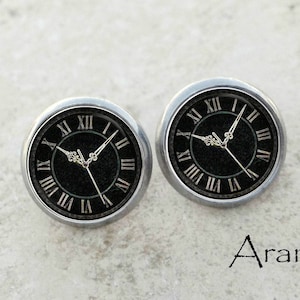 May include: A pair of silver-toned earrings with a black clock face design. The clock face has Roman numerals and a black background.