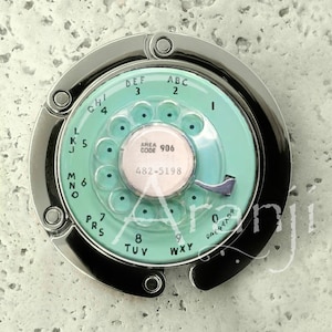 May include: A silver-toned purse hook with a mint green rotary dial design. The dial features numbers, letters, and the text "AREA CODE 906 482-5198". The hook is in a horseshoe shape.